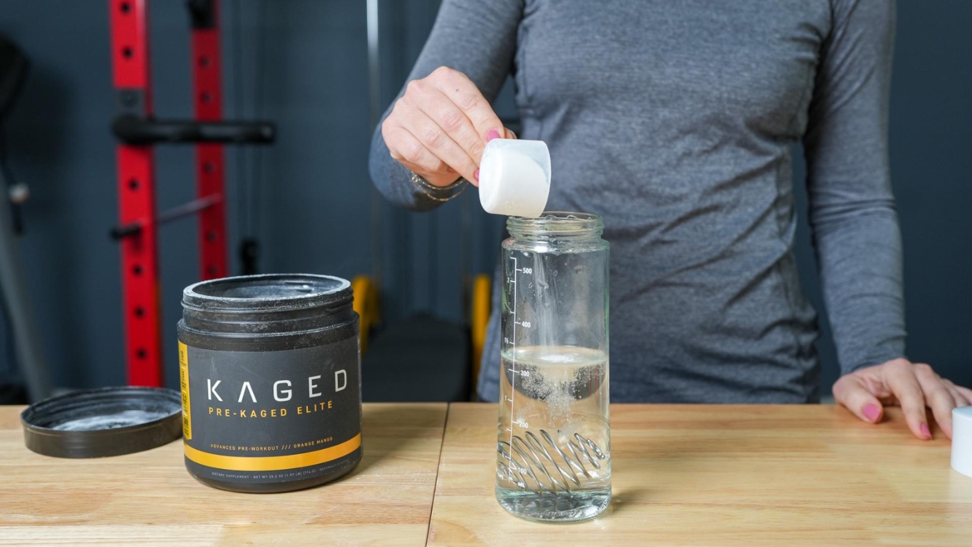 A woman in a gym pouring a scoop of Kaged Pre-Kaged Elite pre-workout powder into a shaker bottle filled with water